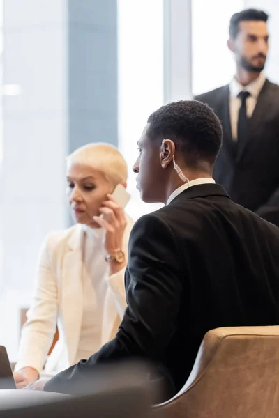 Mujer de negocios borrosa hablando en el teléfono inteligente cerca de guardaespaldas interracial en el vestíbulo del hotel - foto de stock
