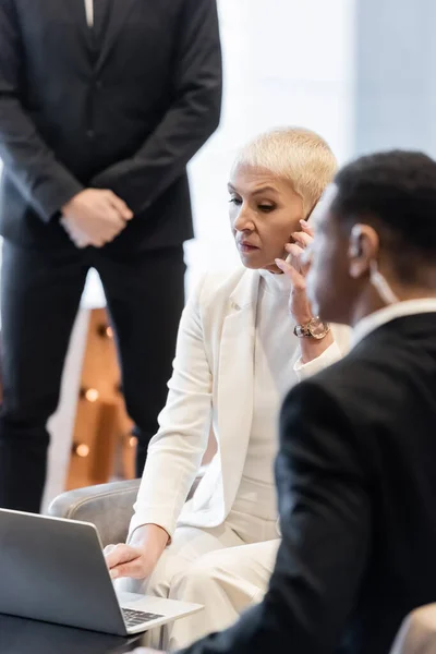 Mujer de negocios hablando por teléfono móvil cerca de la computadora portátil y guardias multiétnicos en el salón del hotel - foto de stock