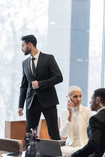 Guardaespaldas bi-racial mirando en la ventana cerca de mujer de negocios madura hablando por teléfono celular en el salón del hotel - foto de stock