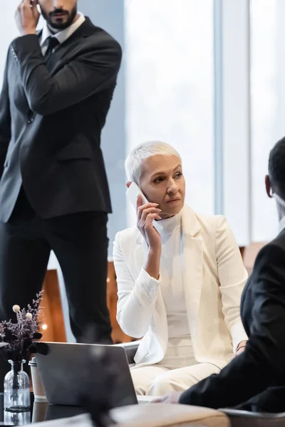 Hombres de seguridad multiétnicos que protegen a la señora mayor de negocios hablando por teléfono celular en el hotel - foto de stock