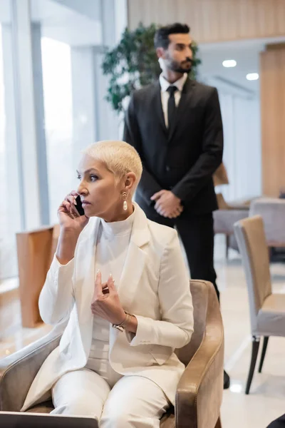 Elegante mujer de negocios senior hablando en el teléfono inteligente en el salón del hotel cerca borrosa guardaespaldas bi-racial - foto de stock