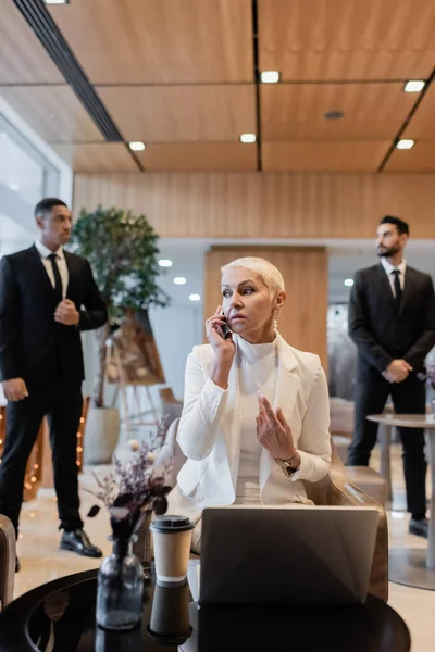 Senior businesswoman talking on smartphone near laptop and blurred multiethnic bodyguards — Stock Photo