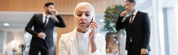 Mature businesswoman talking on smartphone near blurred interracial bodyguards in hotel lounge, banner — Stock Photo