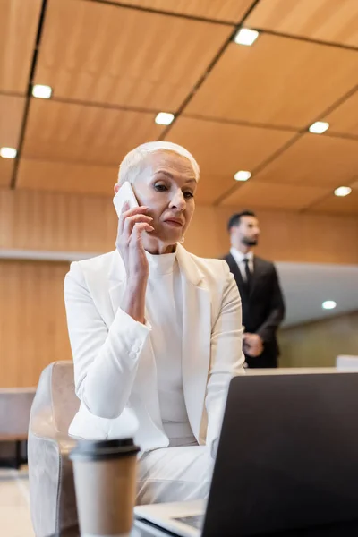 Senior business lady talking on smartphone near blurred laptop and bi-racial bodyguard on background — Stock Photo