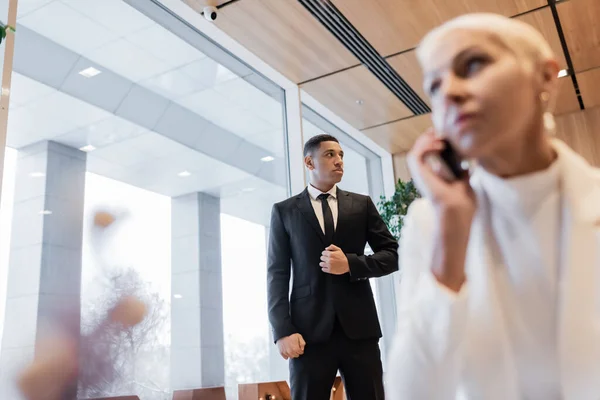 Mujer de negocios borrosa hablando en el teléfono inteligente cerca de guardaespaldas afroamericano en el hotel - foto de stock