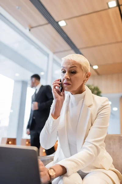 Mature business lady talking on smartphone near laptop and blurred african american security man in hotel lounge — Stock Photo