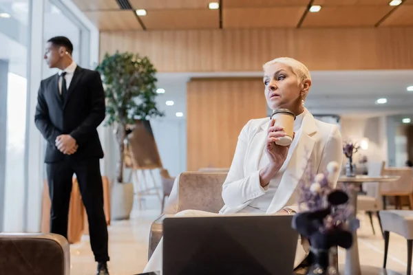 Stylish senior businesswoman with coffee to go sitting near laptop and blurred african bodyguard in hotel lounge — Stock Photo