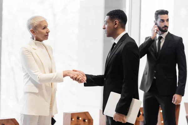 Homem de segurança afro-americano com laptop apertando as mãos com mulher sênior elegante perto de guarda de raça mista — Fotografia de Stock