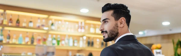 Sonriente hombre de seguridad bi-racial mirando hacia otro lado cerca borrosa bar del vestíbulo en el hotel, pancarta - foto de stock