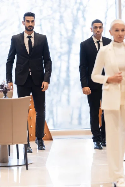 Gardes du corps multiethniques en costumes marchant dans le foyer de l'hôtel près d'une femme d'affaires âgée au premier plan flou — Photo de stock