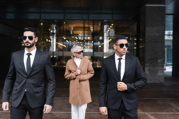 Багатонаціональні охоронці в сонцезахисних окулярах супроводу зрілої бізнес-леді біля входу в готель — стокове фото