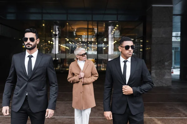 Guardaespaldas multiétnicos en gafas de sol escoltando a mujer de negocios madura cerca de la entrada del hotel - foto de stock