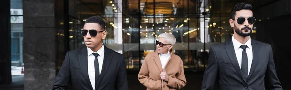 Trendige Senior Business Lady, die auf der Straße in der Nähe von interrassischen Leibwächtern wegschaut, Banner — Stockfoto