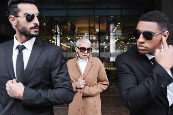 Interracial bodyguards in sunglasses escorting senior business lady near hotel entrance — Stock Photo