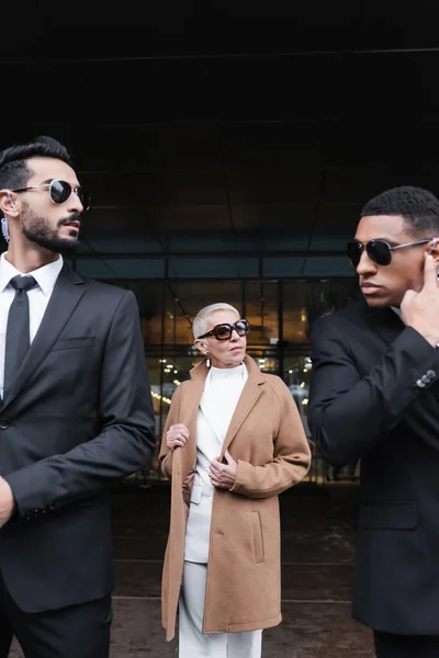 Mujer de negocios senior en abrigo beige y gafas de sol cerca de guardaespaldas privados en la calle urbana - foto de stock