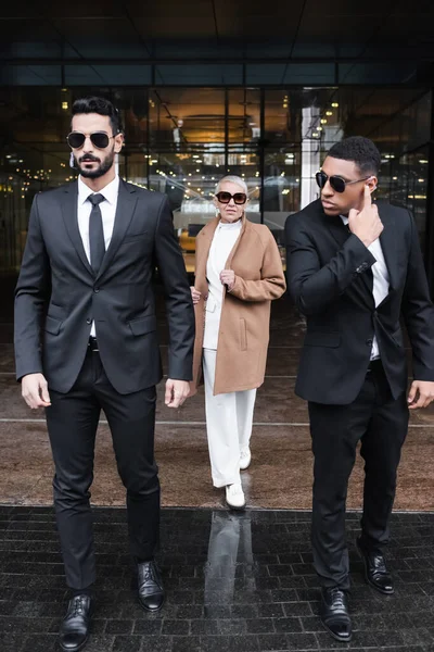 Interracial bodyguards in sunglasses escorting stylish businesswoman near hotel building — Stock Photo