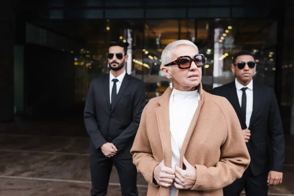 Senior businesswoman in stylish sunglasses looking away near interracial bodyguards outdoors — Stock Photo