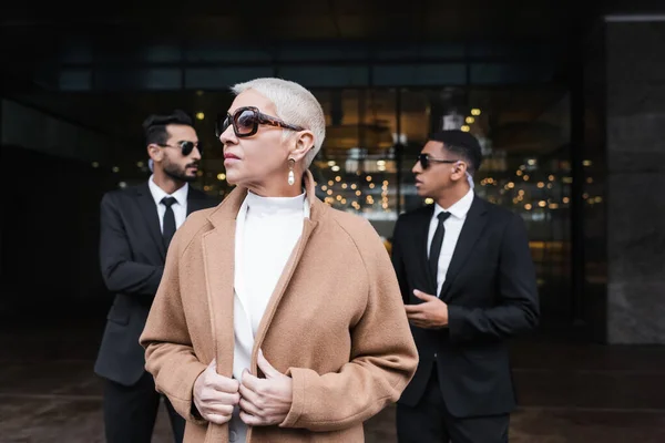 Wealthy businesswoman looking away near hotel entrance and private interracial bodyguards on background — Stock Photo