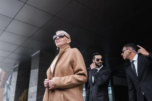 Low angle view of senior businesswoman in trendy coat and sunglasses near interracial bodyguards outdoors — Stock Photo