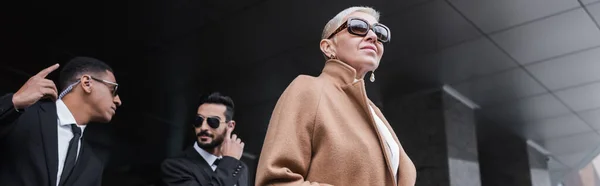 Low angle view of trendy business lady near multiethnic security men outdoors, banner — Stock Photo