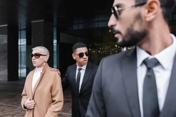 Interracial bodyguards protecting mature business lady near hotel building, blurred foreground — Stock Photo