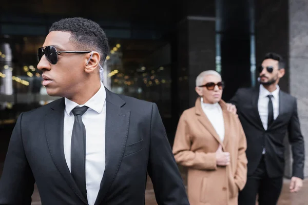 Afro-américain homme de sécurité dans des lunettes de soleil regardant loin près floue femme d'affaires avec garde bi-raciale — Photo de stock