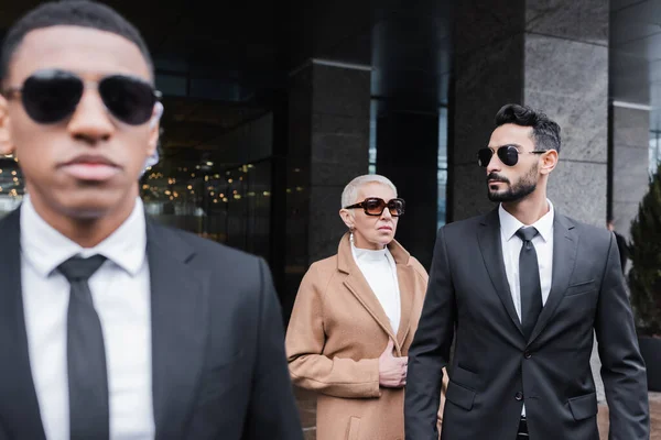 Hombres de seguridad multiétnica en gafas de sol que protegen a la señora madura de negocios cerca del edificio del hotel - foto de stock