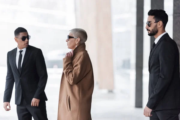 Guardias interracial en trajes negros que protegen elegante mujer de negocios senior en la calle - foto de stock