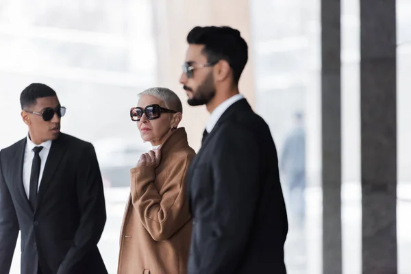 Blurred multiethnic bodyguards escorting stylish senior businesswoman in city — Stock Photo