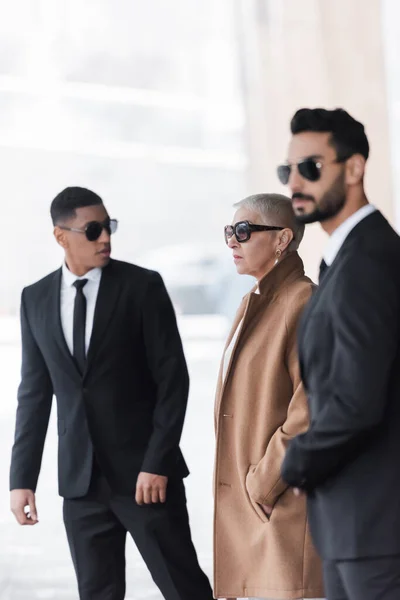 Selective focus of wealthy businesswoman with hand in pocket near private interracial bodyguards outdoors — Stock Photo