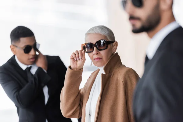 Mature businesswoman adjusting stylish sunglasses near blurred interracial bodyguards on urban street — Stock Photo