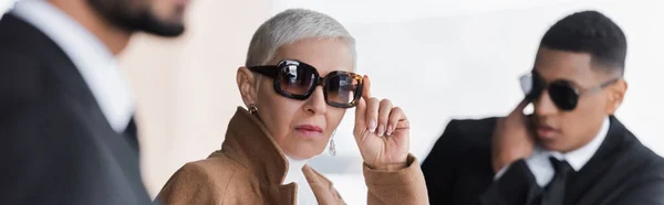 Elegante dama de negocios ajustando gafas de sol cerca borrosa guardaespaldas multiétnicos al aire libre, pancarta - foto de stock