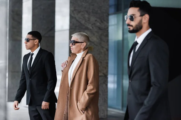 Vista lateral de elegante mujer de negocios con la mano en el bolsillo cerca de guardaespaldas privados interracial en la calle - foto de stock