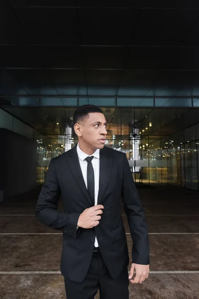 Hombre afroamericano en desgaste formal mirando hacia otro lado mientras está de pie cerca de edificio moderno - foto de stock