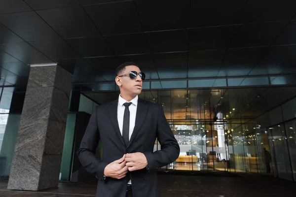 Guardia afroamericana en traje negro y gafas de sol oscuras mirando hacia otro lado cerca del edificio del hotel — Stock Photo