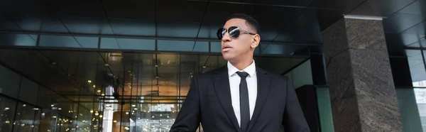 African american security man in formal wear and black sunglasses looking away near hotel entrance, banner — Stock Photo