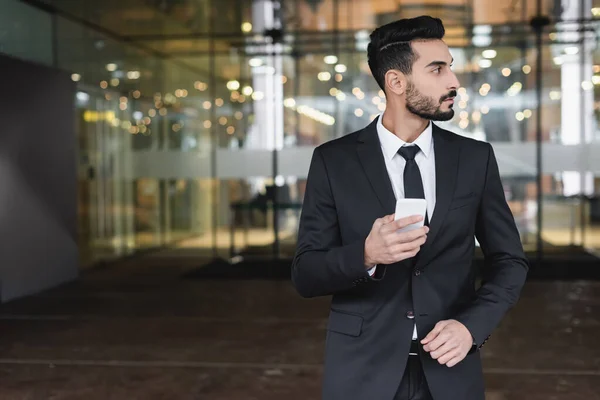 Zwei-Rassen-Sicherheitsmann im Anzug hält Smartphone in der Nähe des Hotelgebäudes und schaut weg — Stockfoto