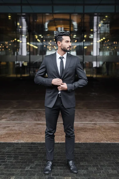 Full length view of mixed race guard buttoning blazer and looking away near blurred hotel entrance — Stock Photo