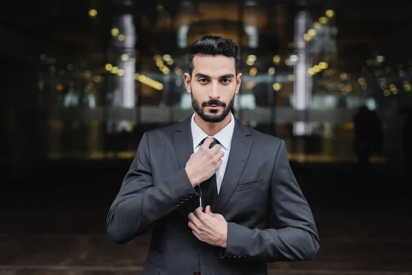 Bearded mixed race security man looking at camera and adjusting tie outdoors — Stock Photo