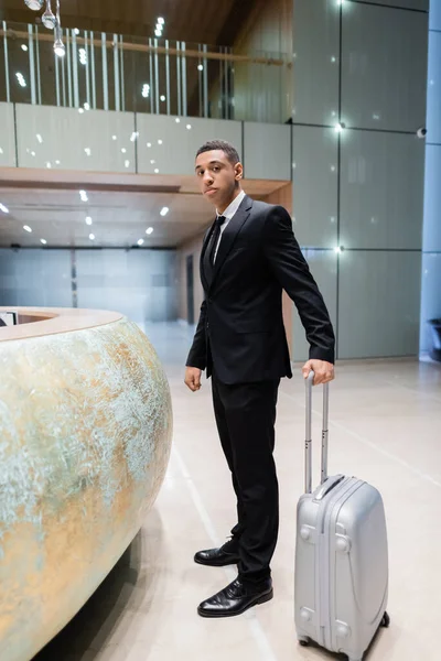 Volle Länge Ansicht von afrikanisch-amerikanischen Sicherheitsmann mit Koffer Blick auf Kamera in der Nähe der Hotelrezeption — Stock Photo