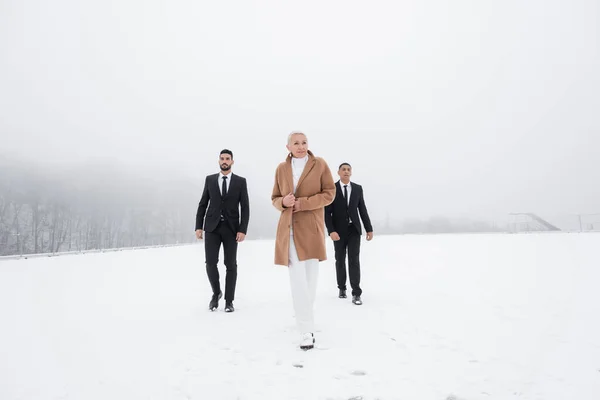 Full length view of stylish senior businesswoman walking across winter field with interracial bodyguards — Stock Photo