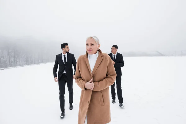 Empresária sênior em casaco andando através do campo de inverno perto de guarda-costas multiétnicos em ternos — Fotografia de Stock