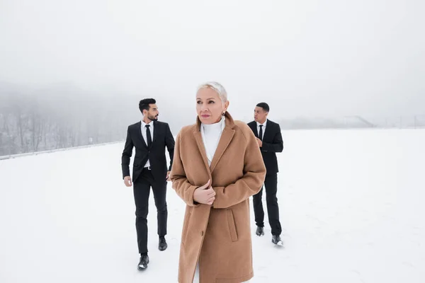 Seniorin im Mantel läuft in Anzug über Winterfeld in der Nähe multiethnischer Leibwächter — Stockfoto