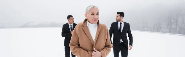 Mature businesswoman in coat near interracial bodyguards on winter field, banner — Stock Photo