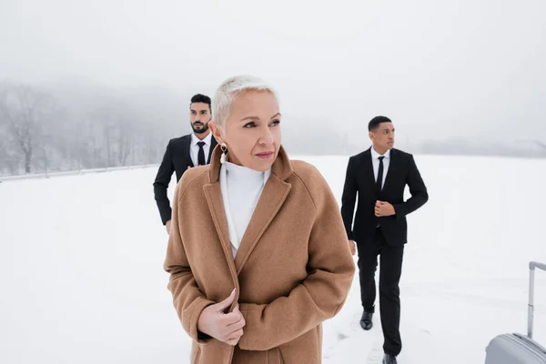 Maduro mujer de negocios mirando lejos cerca de multiétnicos hombres de seguridad en el campo cubierto de nieve - foto de stock