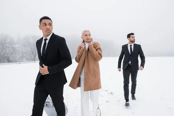 Multicultural guards in black suits protecting senior business lady while walking on winter field — Stock Photo