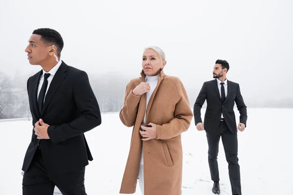 Interracial bodyguards in formal wear walking near senior businesswoman on snowy field — Stock Photo