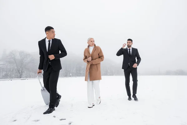 Homem de segurança afro-americano carregando mala perto de empresária sênior e guarda-costas bi-racial no campo nevado — Fotografia de Stock