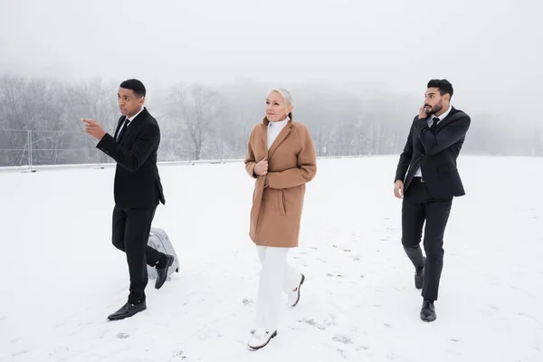 Guardaespaldas afroamericano con maleta apuntando dirección cerca de la mujer de negocios senior en el campo cubierto de nieve - foto de stock