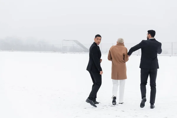 Hombre de seguridad afroamericano mirando hacia atrás mientras camina cerca de la mujer de negocios senior y guardaespaldas bi-racial - foto de stock